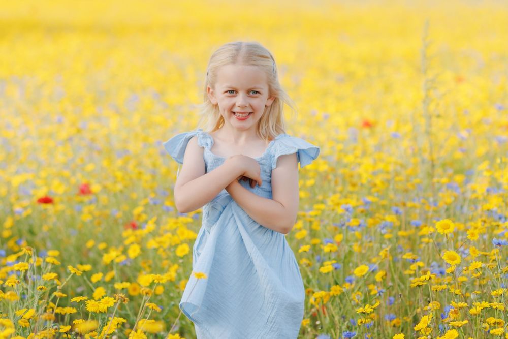 A child in a light blue dress stands joyfully in a vibrant field of blooming yellow wildflowers.