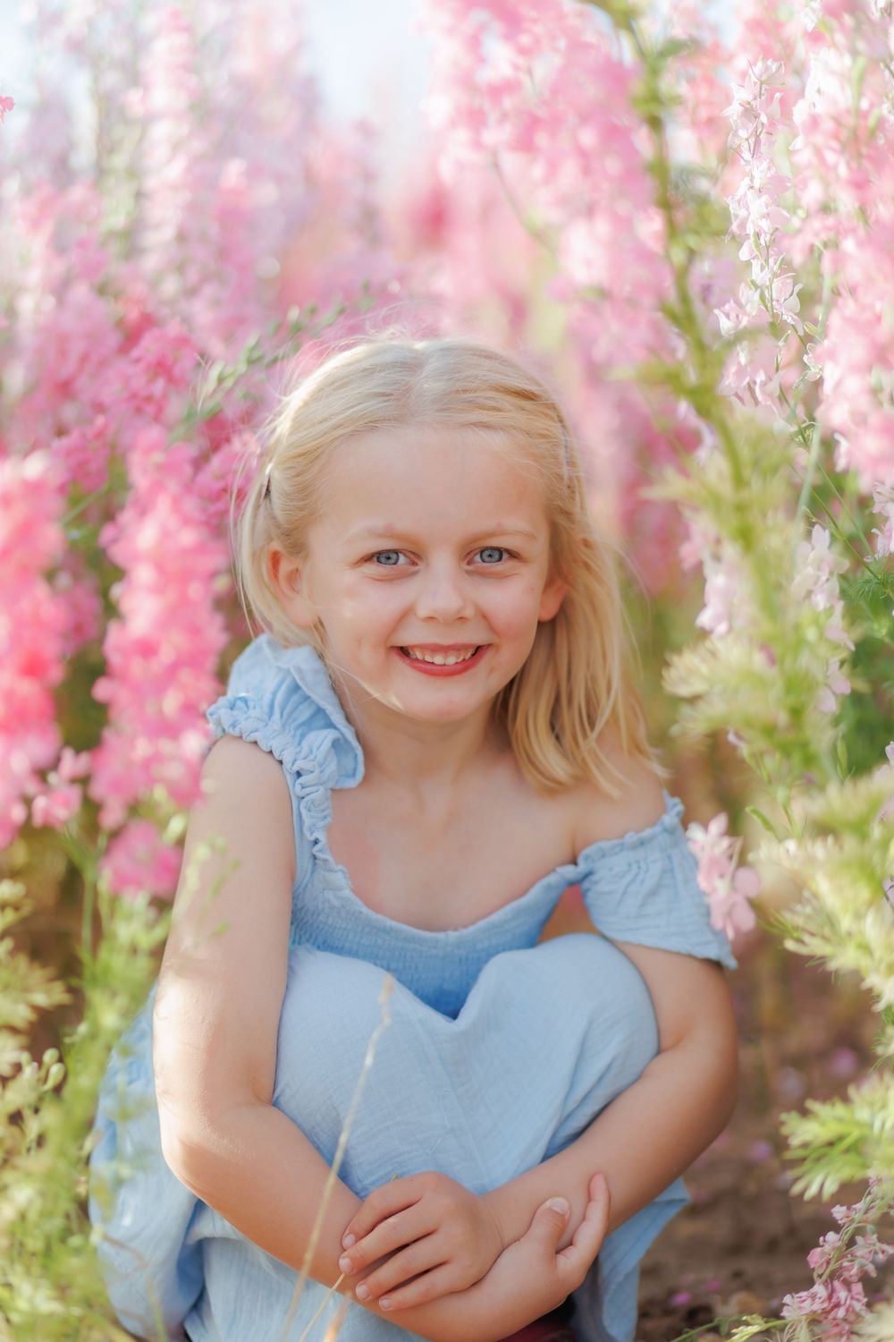 Child in light blue dress poses among pink blossoms during springtime photoshoot in garden.
