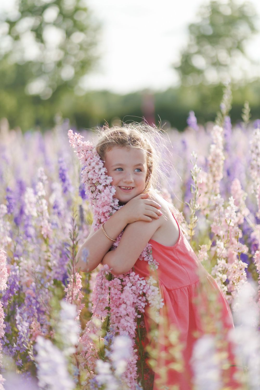A child in a coral dress stands among blooming pink and purple flowers in a summer garden during golden hour.