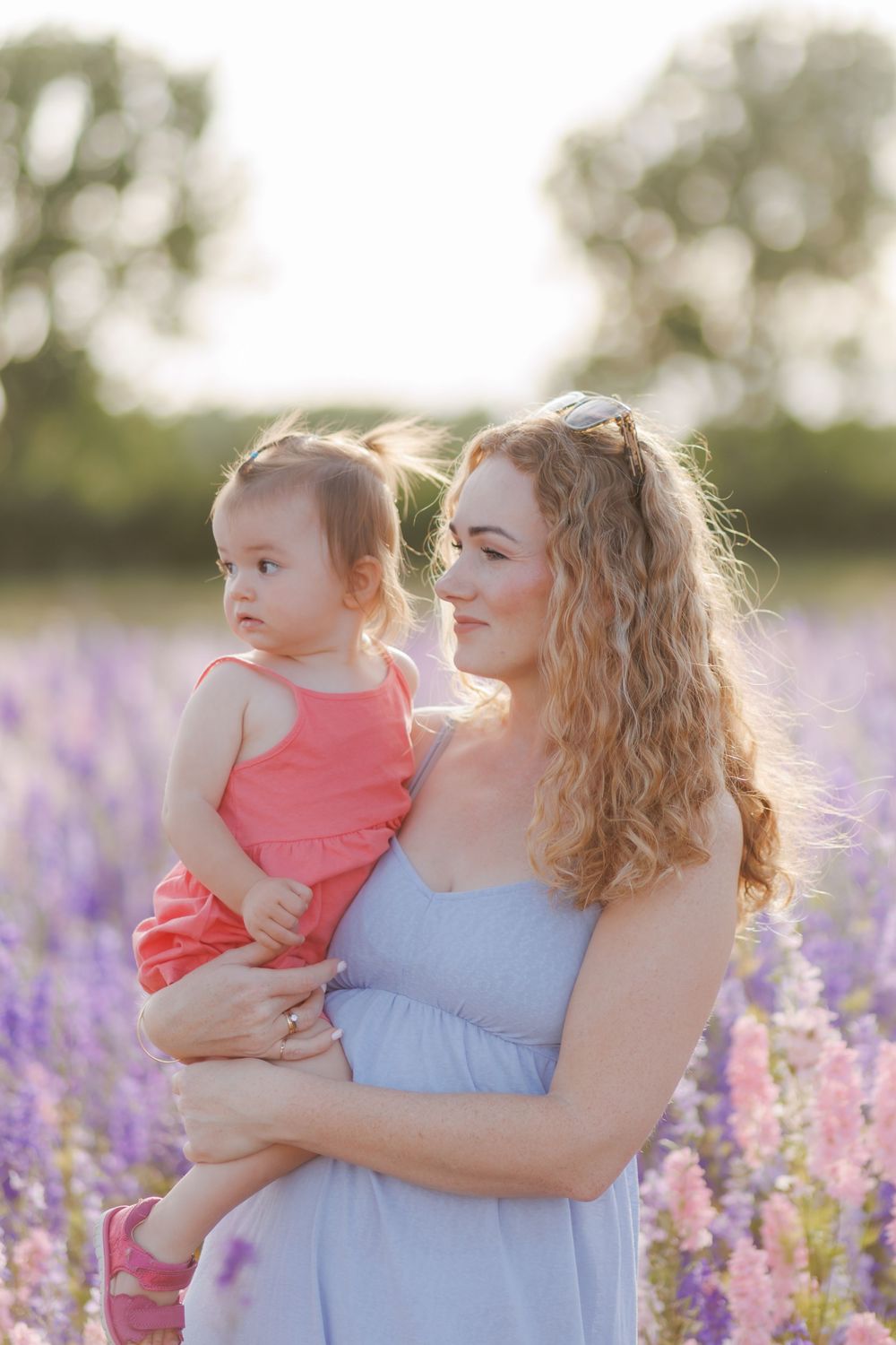 Mother and daughter embrace in a dreamy field of lavender at sunset while wearing coral and blue dresses.