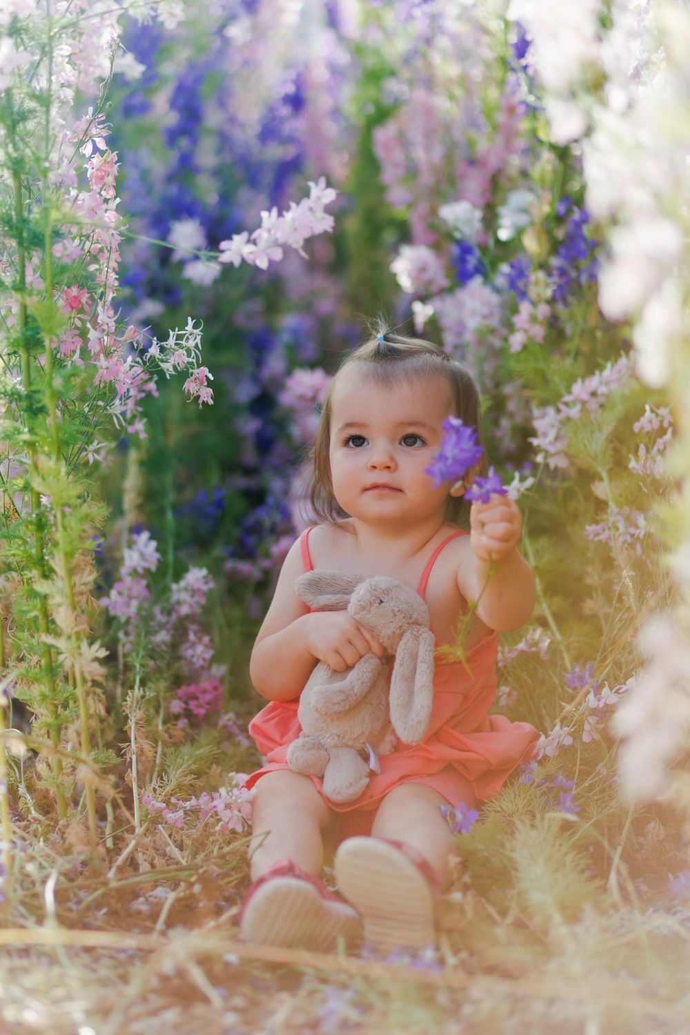A toddler in a pink dress sits among blooming purple and pink wildflowers while holding a plush bunny toy.
