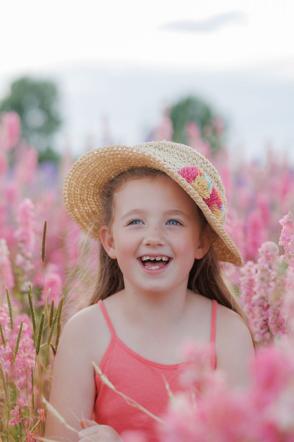 A happy child wearing a straw sunhat and coral dress smiles joyfully among pink wildflowers on a sunny summer day.
