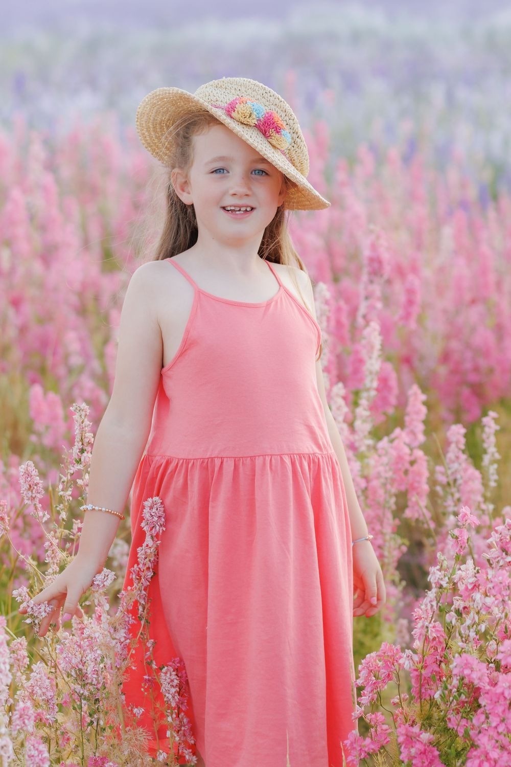 A person in a coral pink sundress and a straw hat stands in a field of blooming pink wildflowers on a summer day.