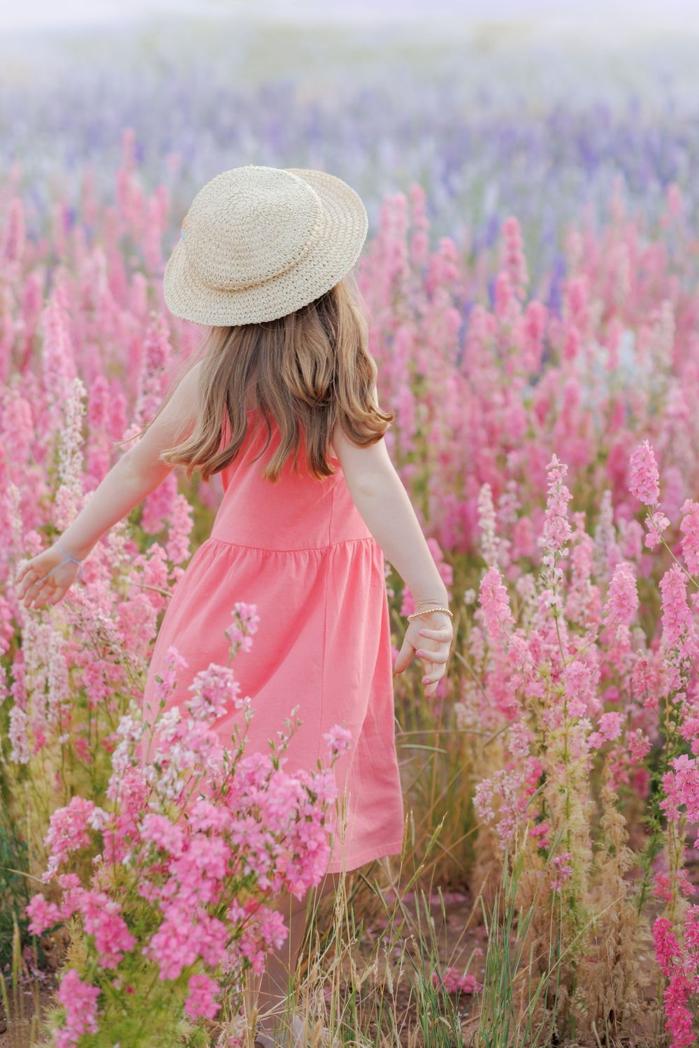 A person in a pink sundress and straw hat walks through a vibrant field of pink and purple wildflowers on a summer day.