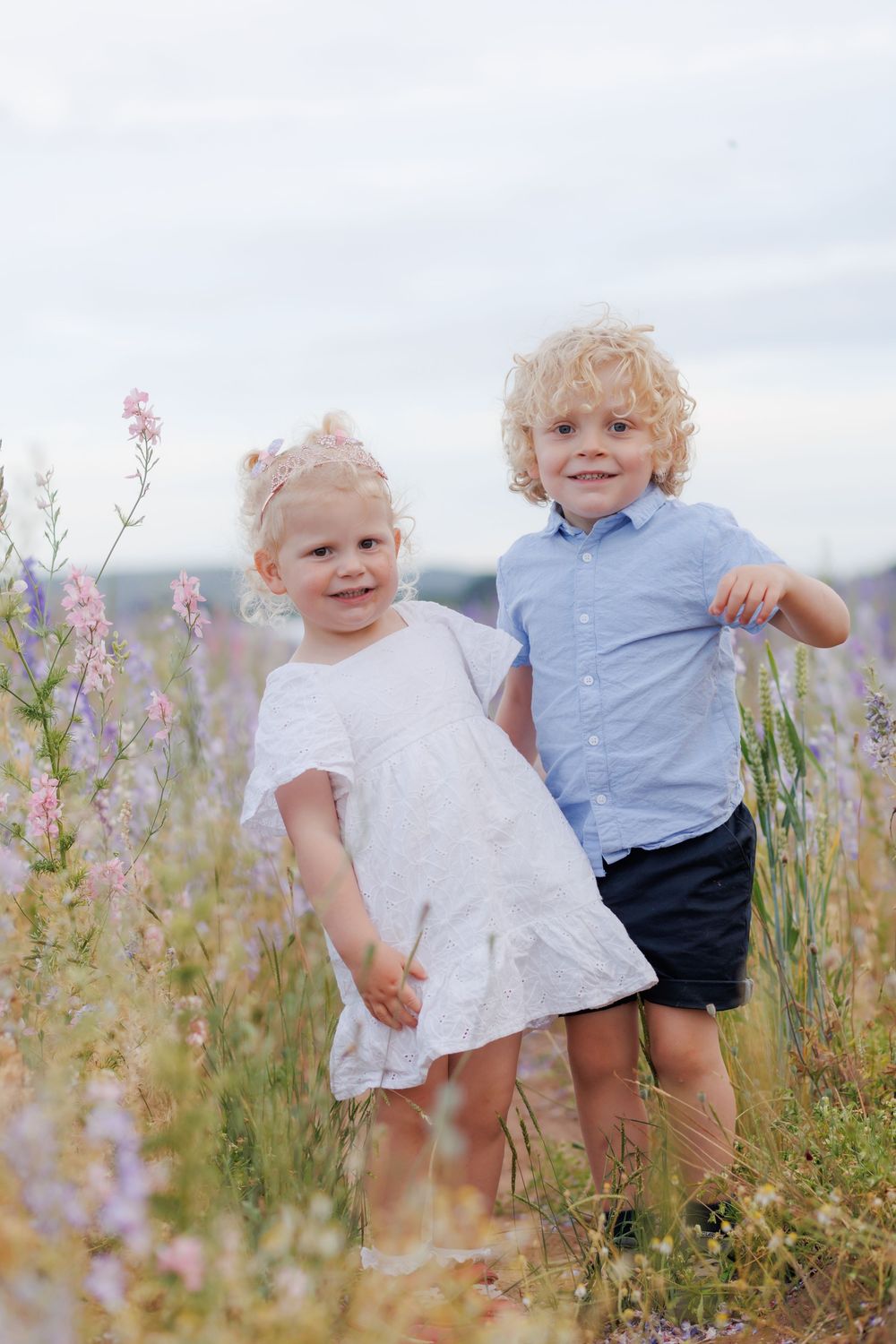 Two children sit among blooming wildflowers in a meadow on a cloudy day.