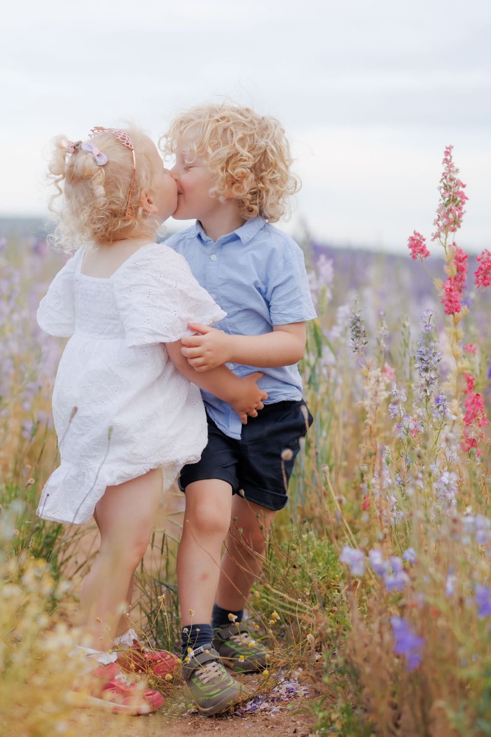 Two curly-haired children share a tender moment in a wildflower meadow filled with lavender and pink blooms.