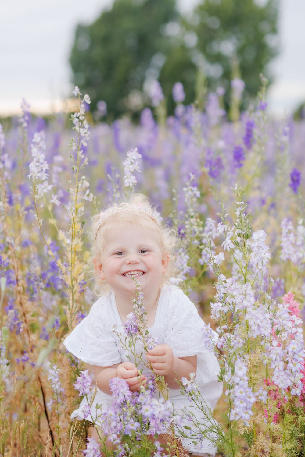 A child wearing white smiles joyfully while sitting among blooming purple and pink wildflowers in a summer meadow.