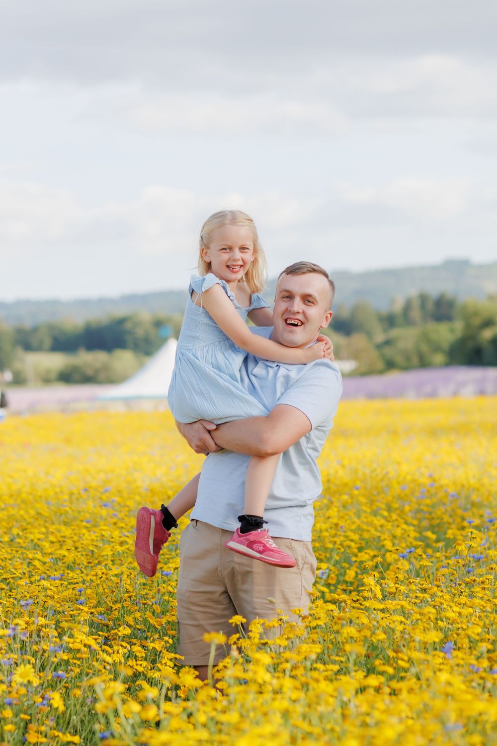 Two figures share a joyful moment in a vibrant yellow wildflower meadow with rolling hills in the background.