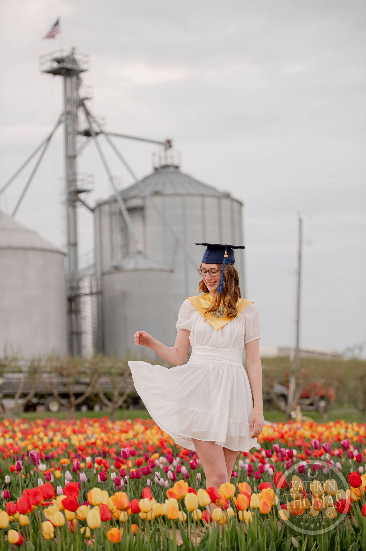 Graduate in cap and gown poses in vibrant tulip field with large grain silos in background on cloudy day.
