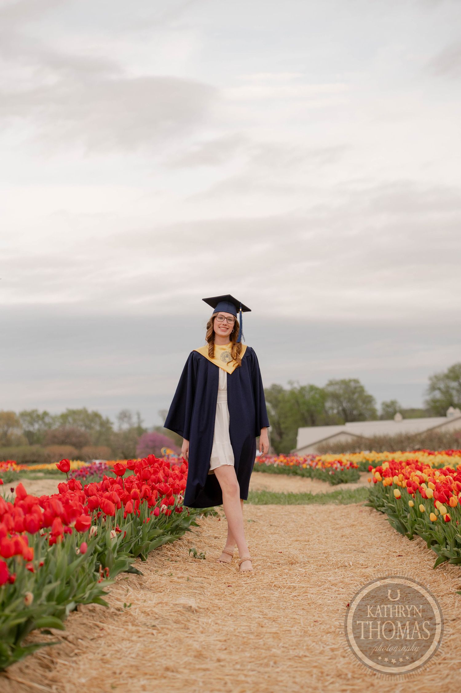 Graduate in cap and gown stands in a pathway surrounded by blooming red tulips on a cloudy spring day.