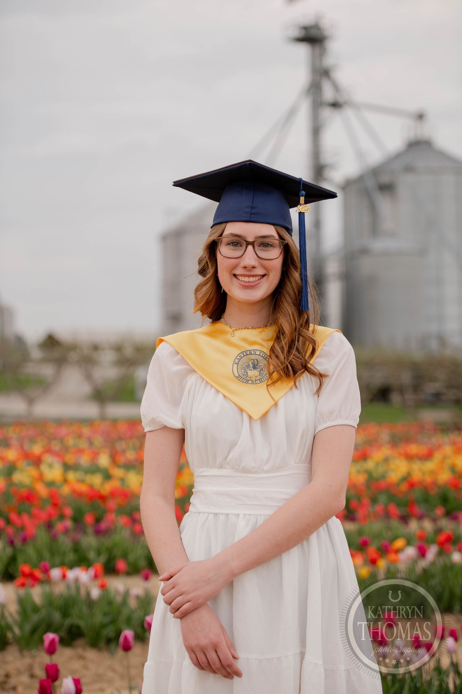 A graduation portrait in a blooming tulip field with white grain silos in the background on an overcast day.