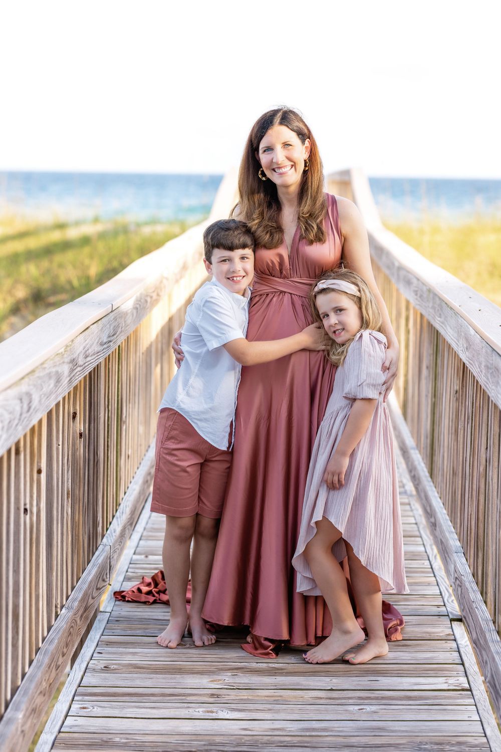 A family stands together on a wooden boardwalk at the beach with the ocean visible in the background.