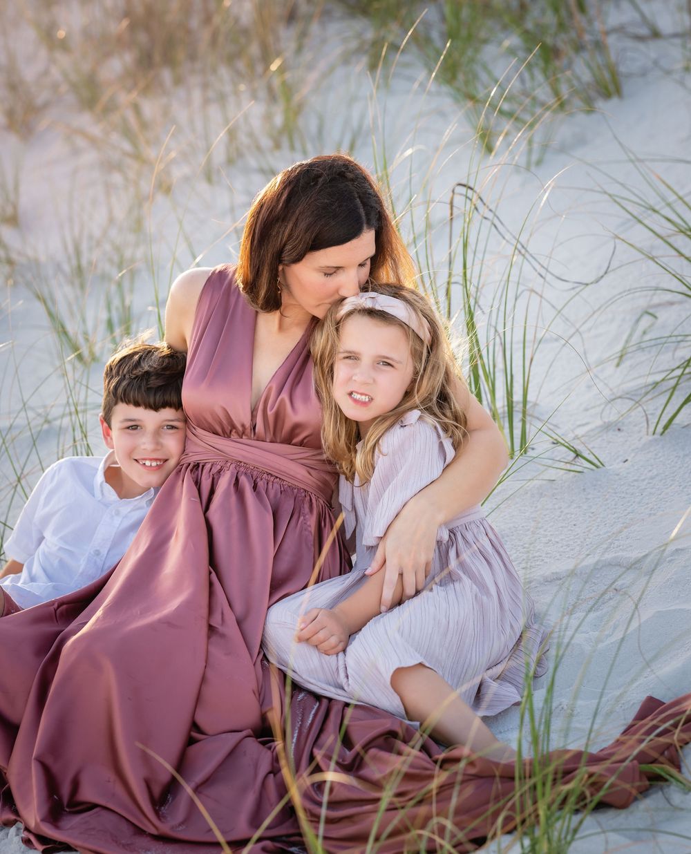 A sweet family moment captured in the dunes with beach grass, wearing mauve and light colored dresses at sunset.