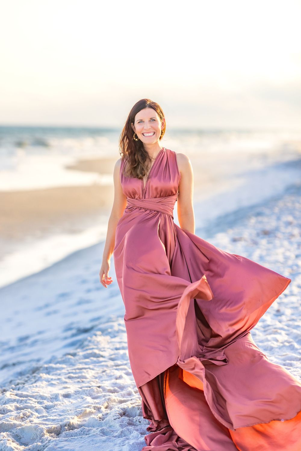 A flowing mauve evening gown catches the wind on a white sandy beach at sunset with ocean waves in the background.