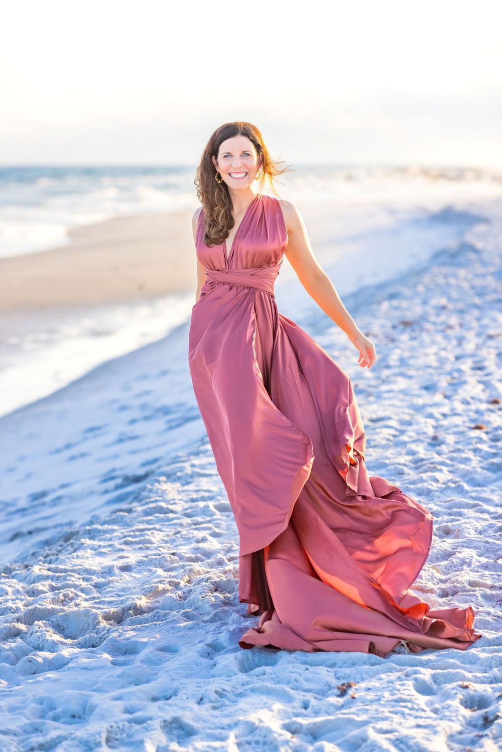 A flowing mauve dress catches the ocean breeze as someone walks along a snowy white beach at sunset.