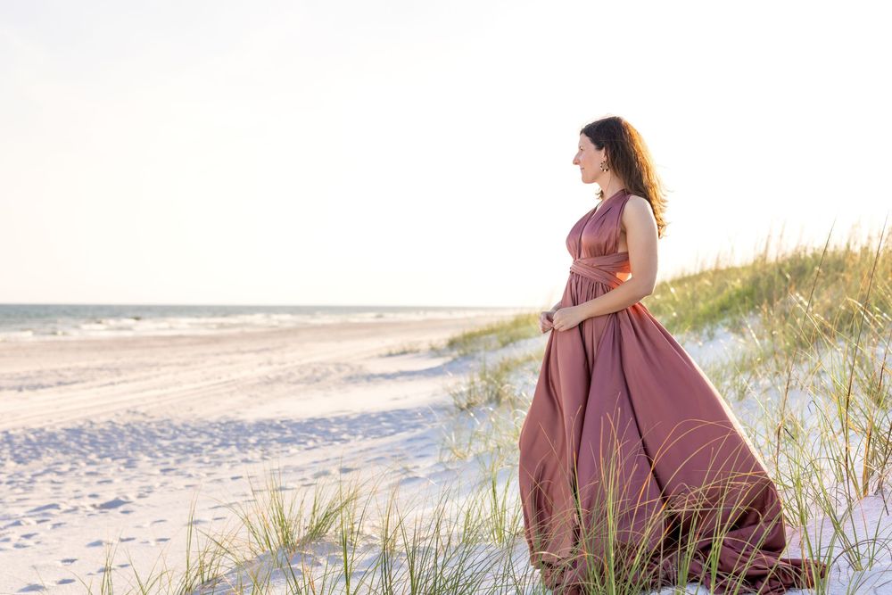A person in a flowing mauve vintage dress stands on a sandy beach with sea grass and ocean waves in the background.