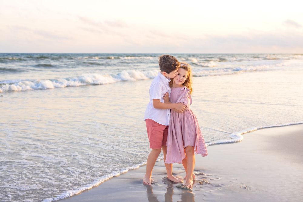 A couple embraces while standing barefoot at the ocean's edge on a sandy beach during sunset.