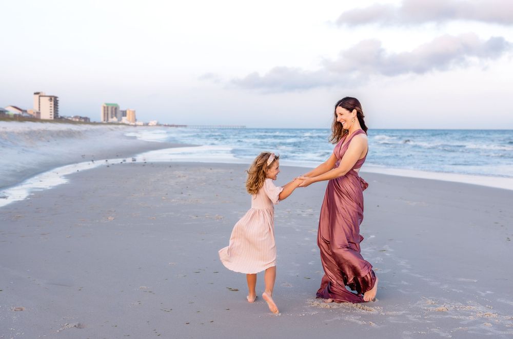Two people in flowing dresses dance together on a beach at sunset with coastal buildings visible in the distance.
