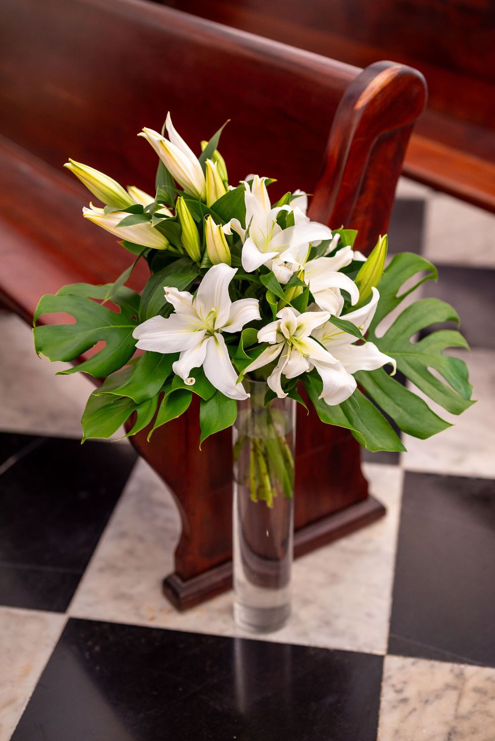 Elegant white lily bouquet with monstera leaves arranged in a glass vase on a checkered floor.