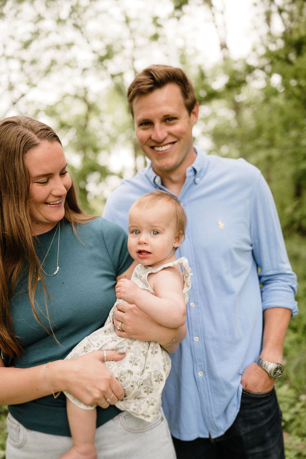 Family portrait outdoors with baby wearing floral dress and parents in casual attire.