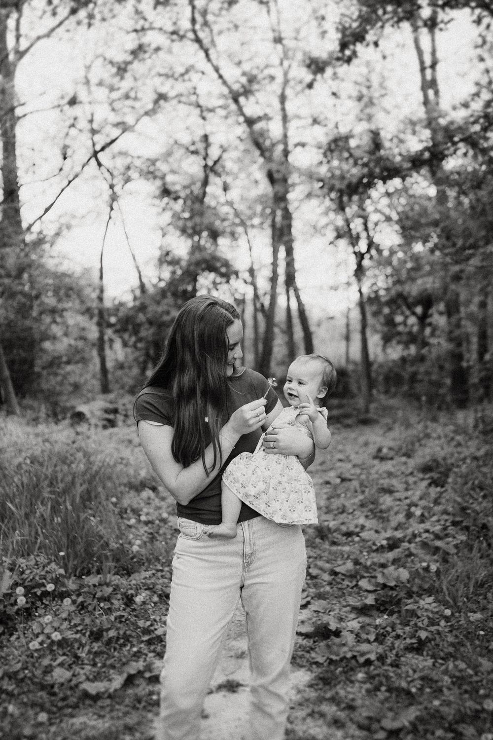 A tender moment captured in black and white of a parent and baby standing together in a wooded forest setting.