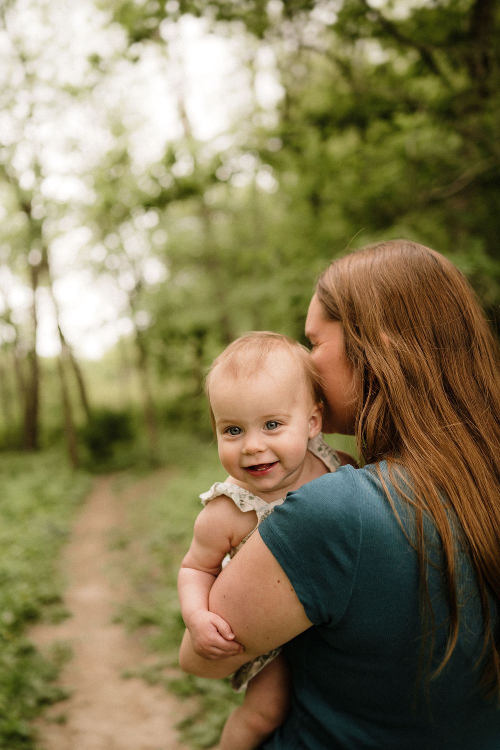 Parent in teal shirt holds and kisses smiling child while standing on nature trail.