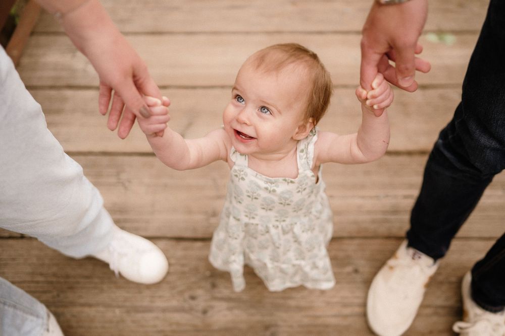 Top view of baby in white dress holding parents' hands on wooden floor.