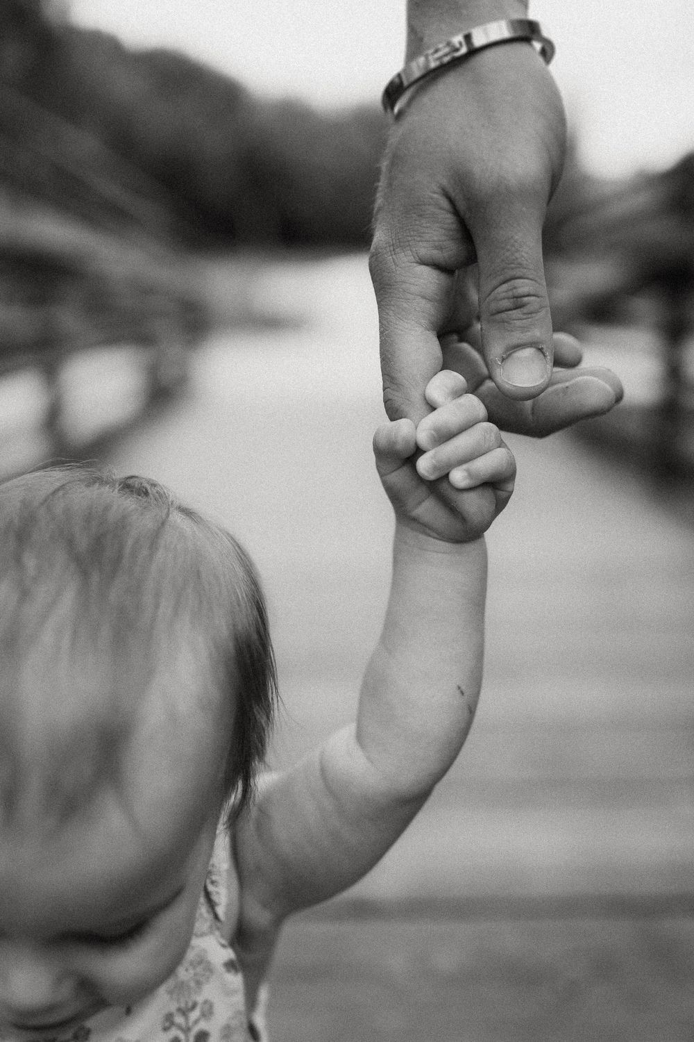 Black and white image showing a small child holding an adult's finger while walking together outdoors.