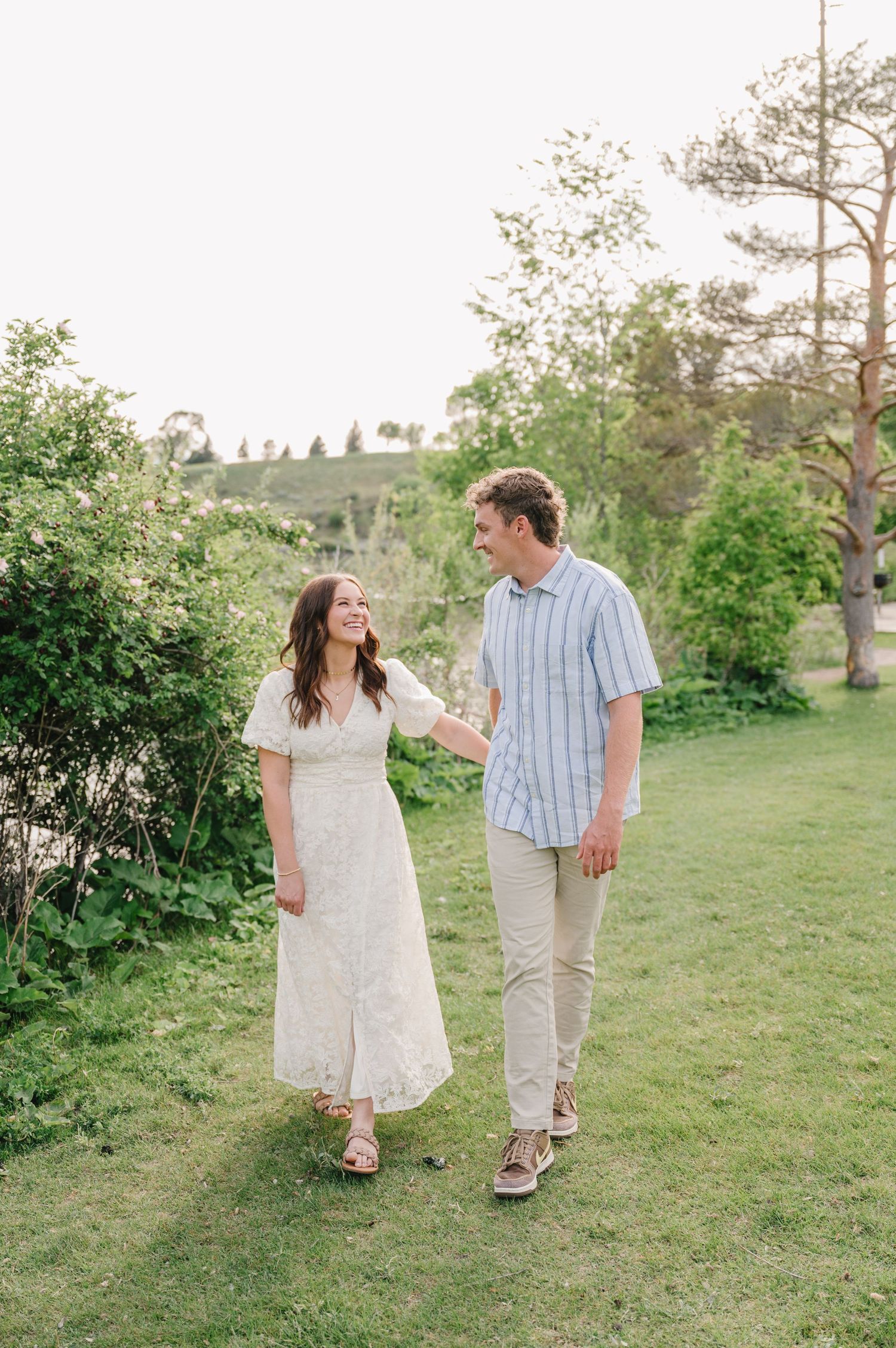 A couple walks together on a lush green lawn surrounded by trees on a sunny summer day.