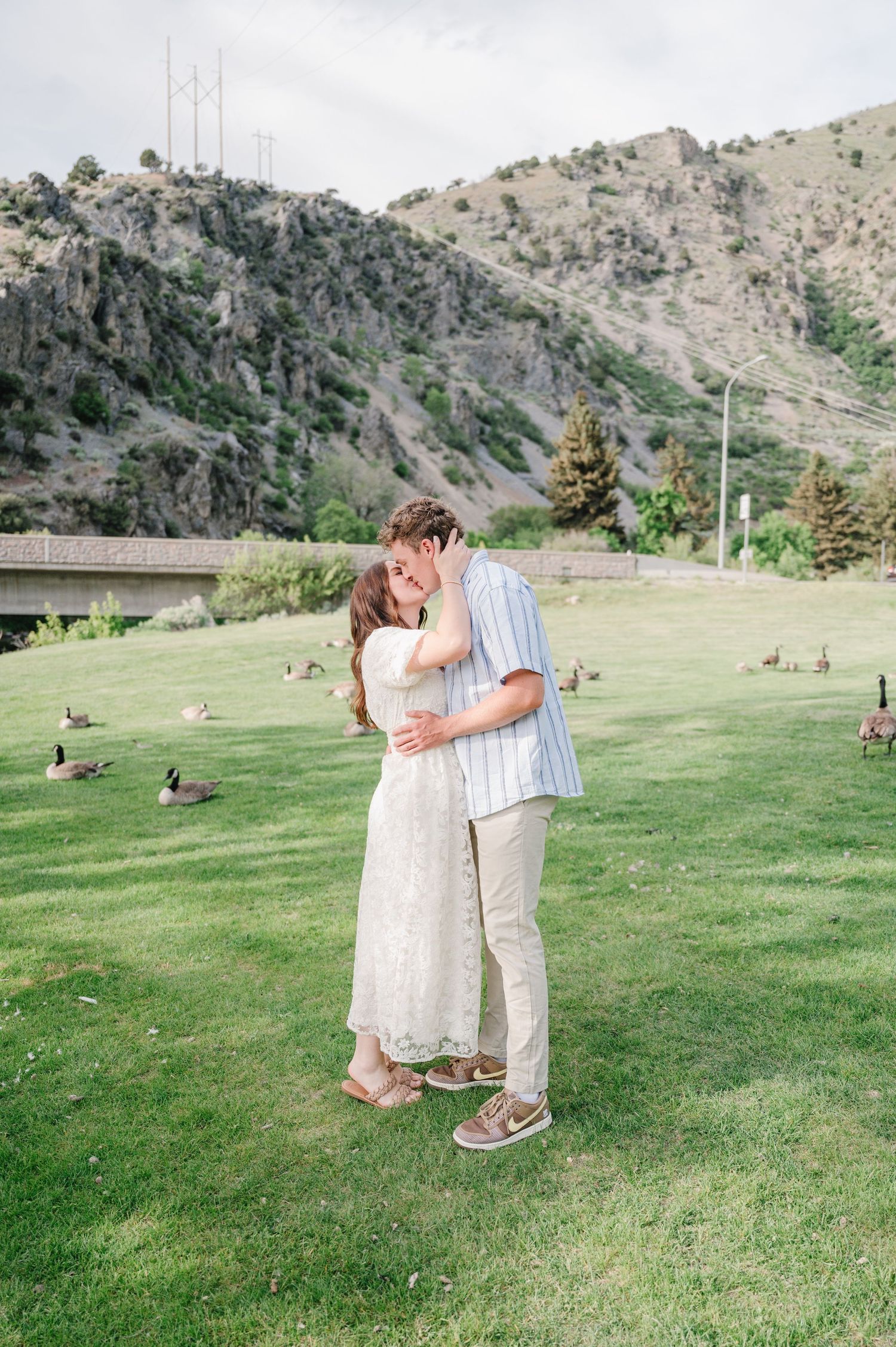 A couple shares a romantic moment on a grassy hill with mountains and transmission towers in the background.
