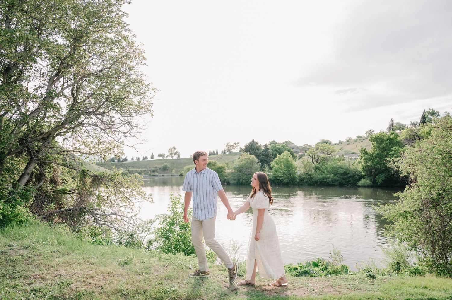 A couple walks hand in hand along a riverbank on a sunny day surrounded by lush greenery and trees.