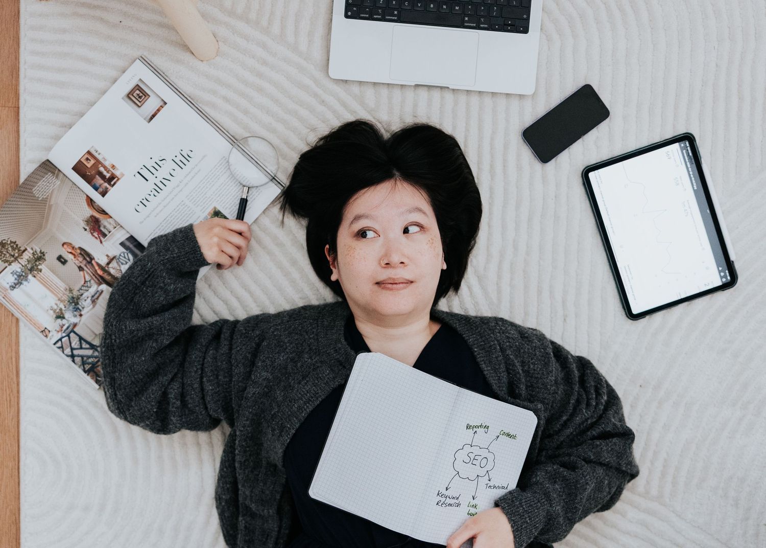 Person in black sweater lying on white surface with scattered papers and digital devices around them.