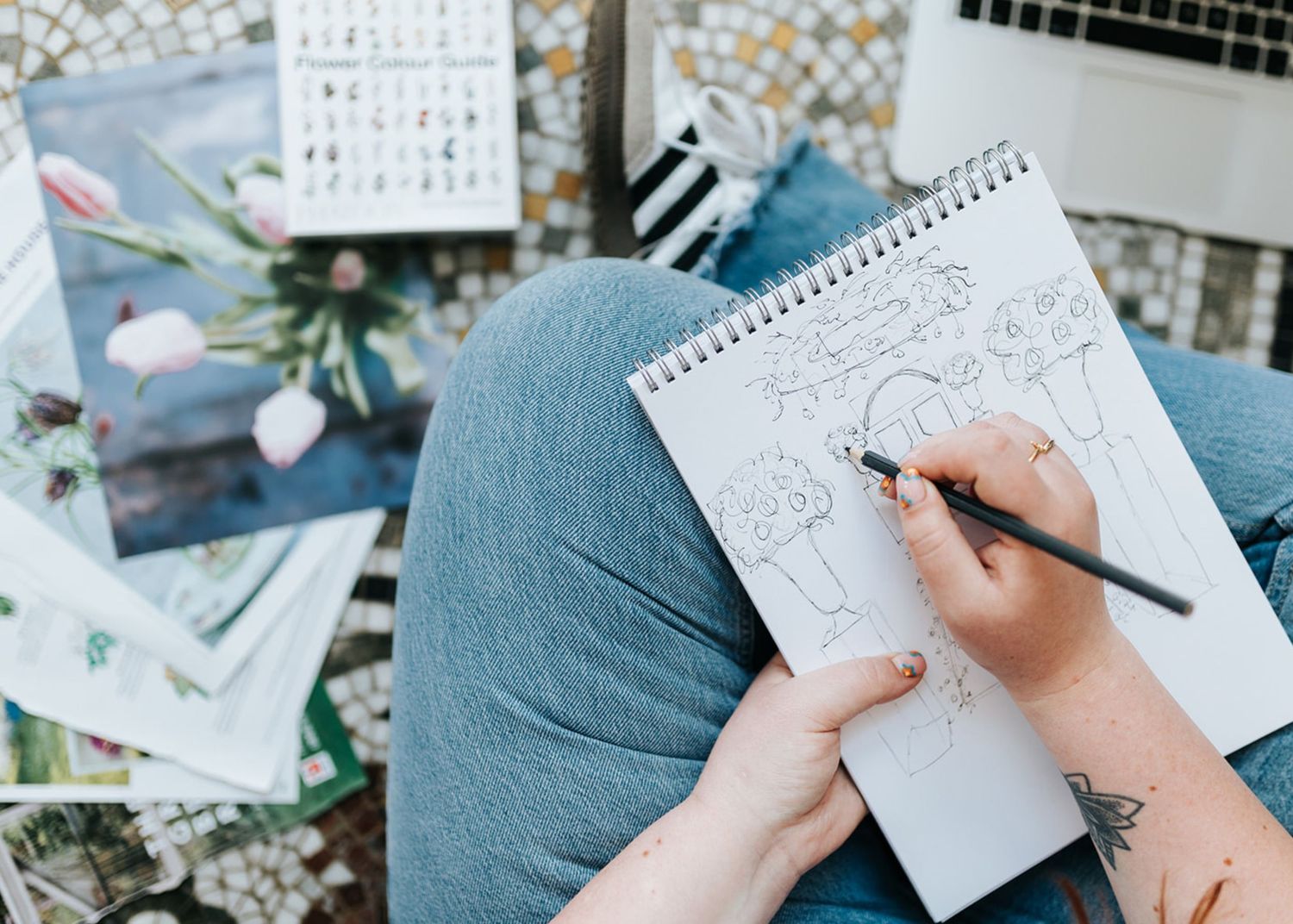 Artist sketching in a notebook while sitting on a blue cushion with reference photos scattered around.