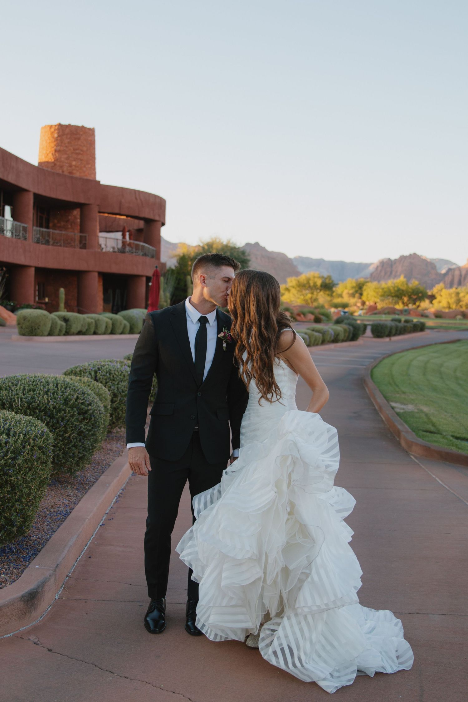 A couple in wedding attire walks hand in hand along a scenic pathway at a desert resort at sunset.