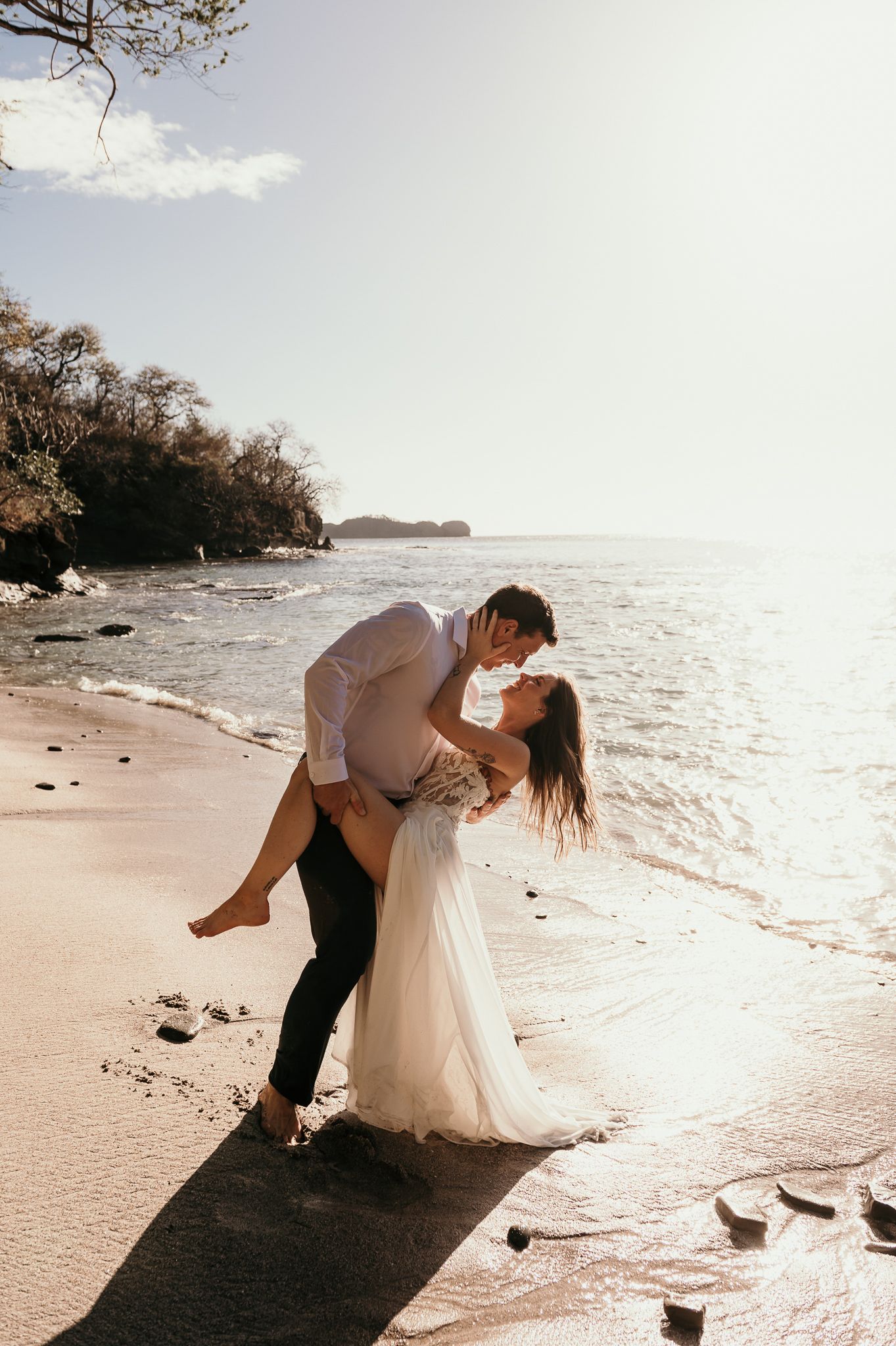 A romantic couple embraces on a serene beach at sunset with waves lapping at the shore and trees in the background.