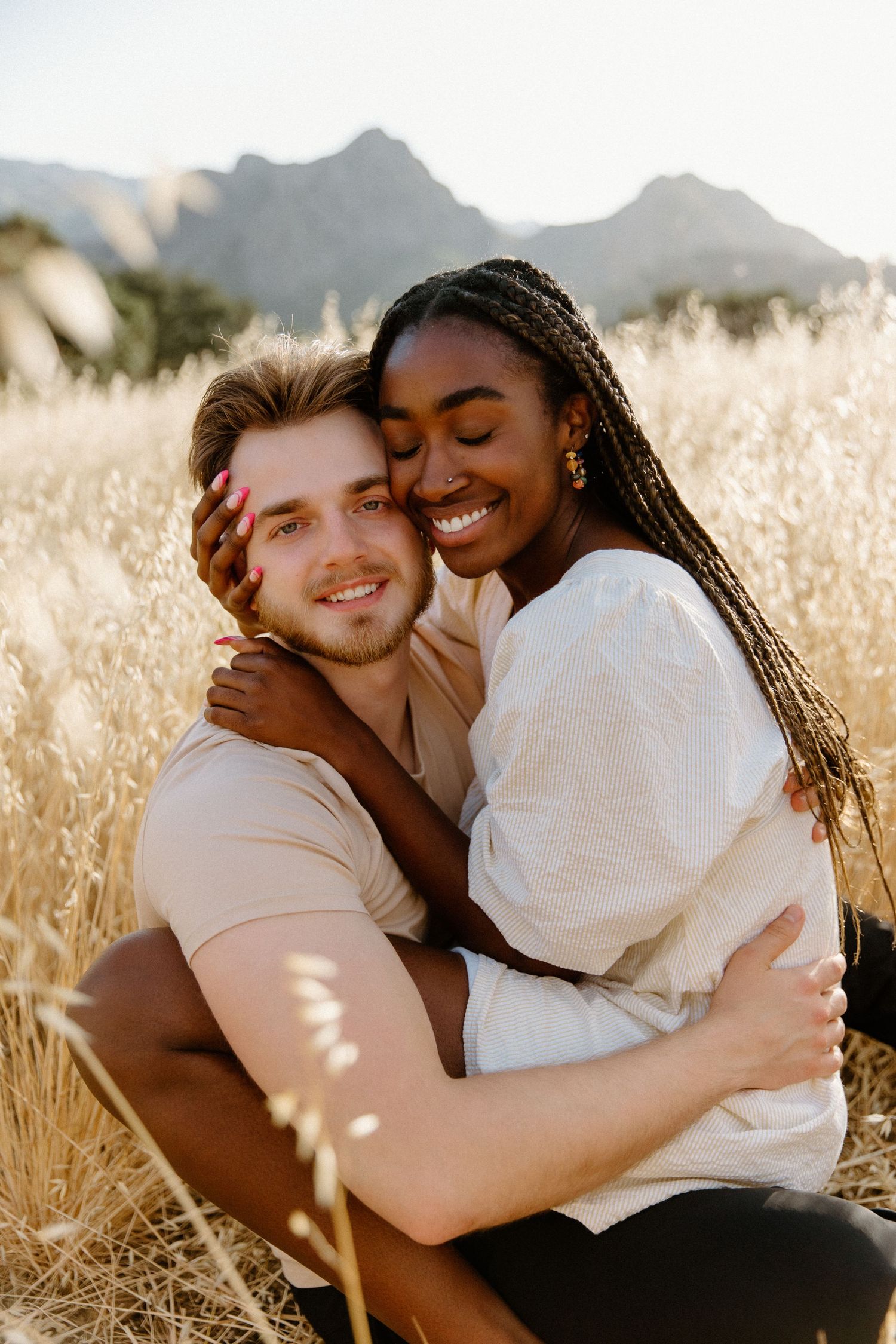 A couple shares an intimate embrace in a golden wheat field with mountains in the background at sunset.