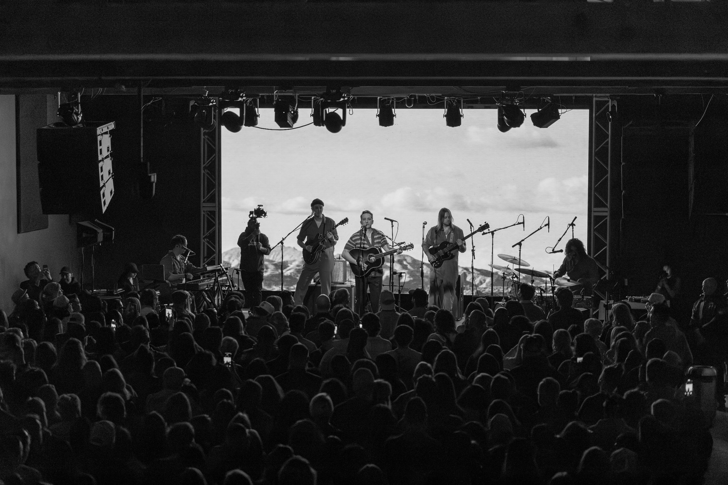 A black and white concert photo showing a band performing on stage in front of a large crowd at a venue.