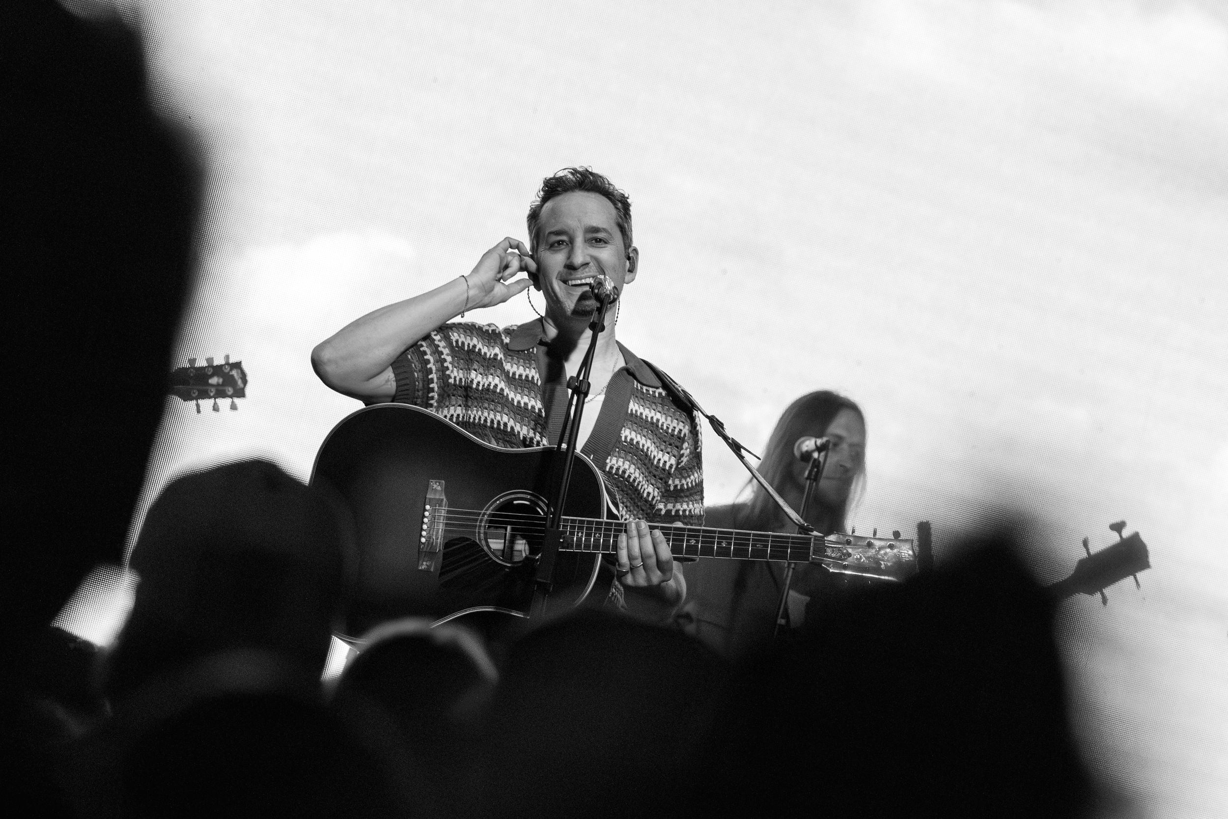 Black and white concert photo of a performer on stage singing and playing acoustic guitar in front of a crowd.