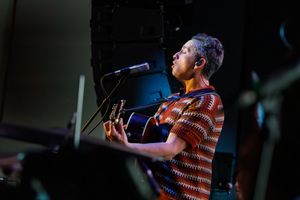 Musician in striped shirt performs on stage with acoustic guitar and microphone in dramatic lighting.