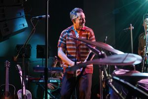 Musician in a striped shirt performs on stage with drum kit and musical equipment under blue lighting.