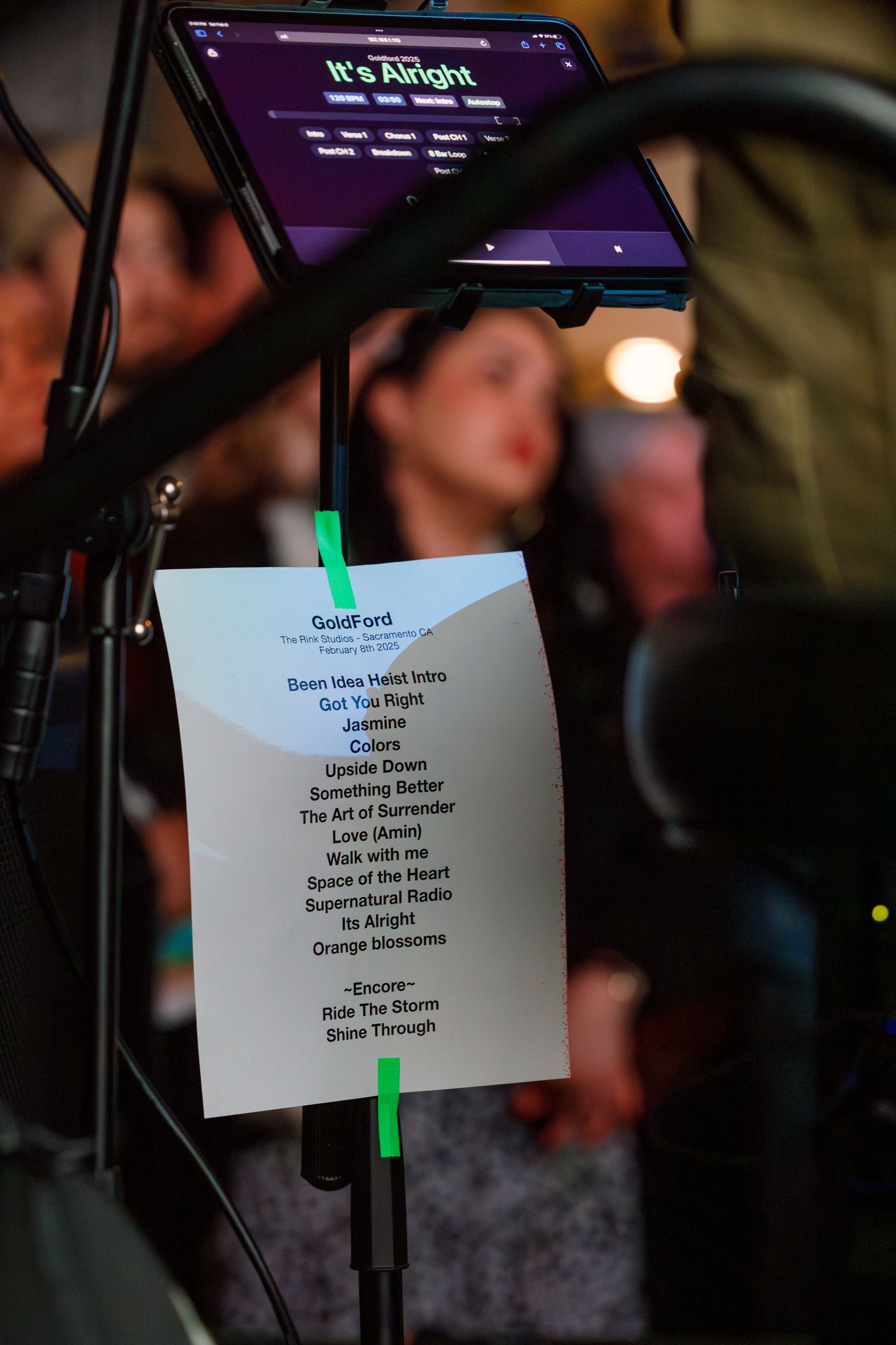 A music setlist clipped to a microphone stand with a digital tablet mounted above it in purple lighting conditions.