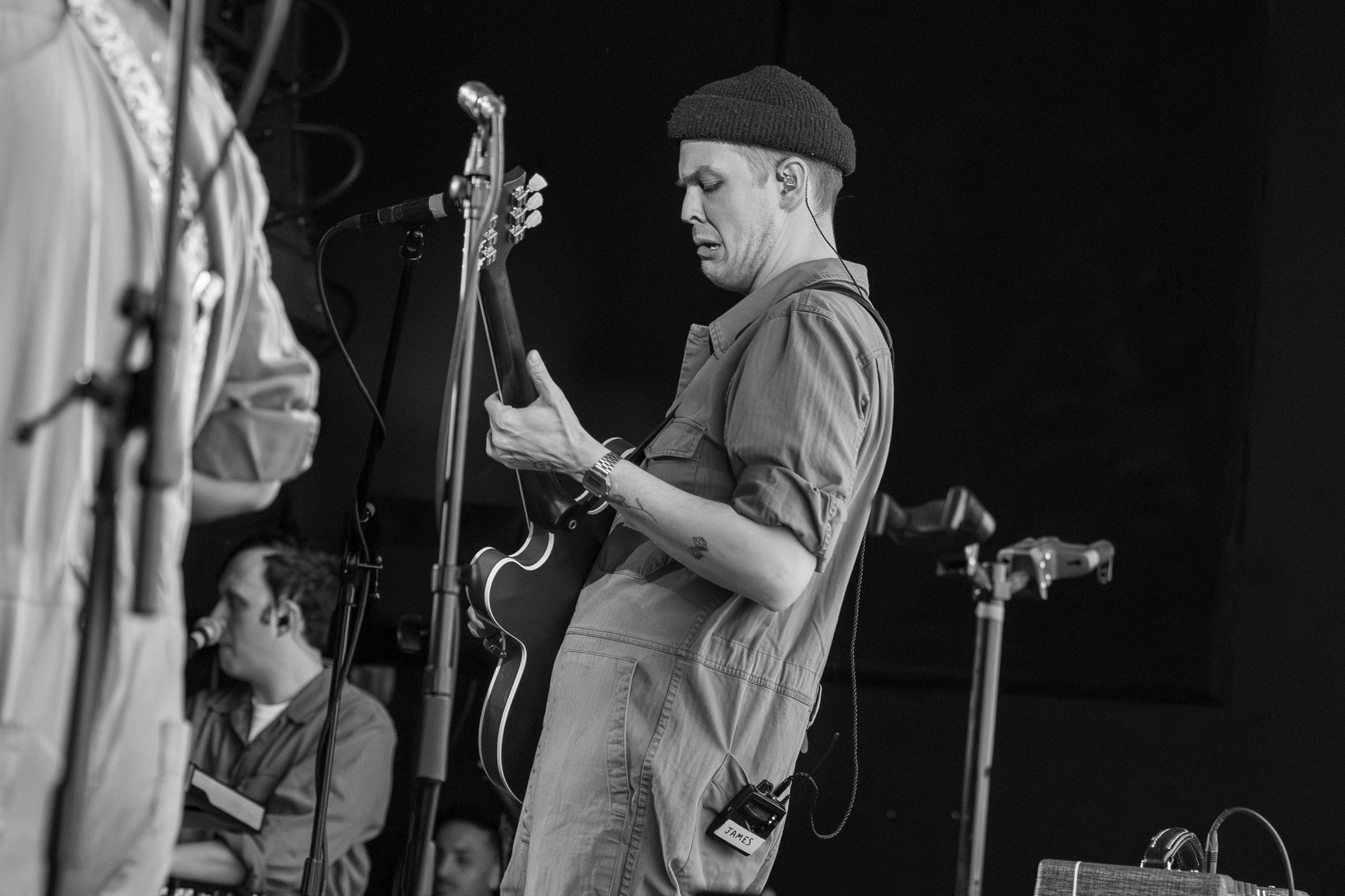 A musician performs on stage wearing a beanie while playing bass guitar during a live performance in black and white.