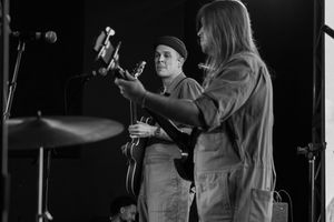 Musicians perform with acoustic guitar while standing near microphones on a dark stage in black and white.