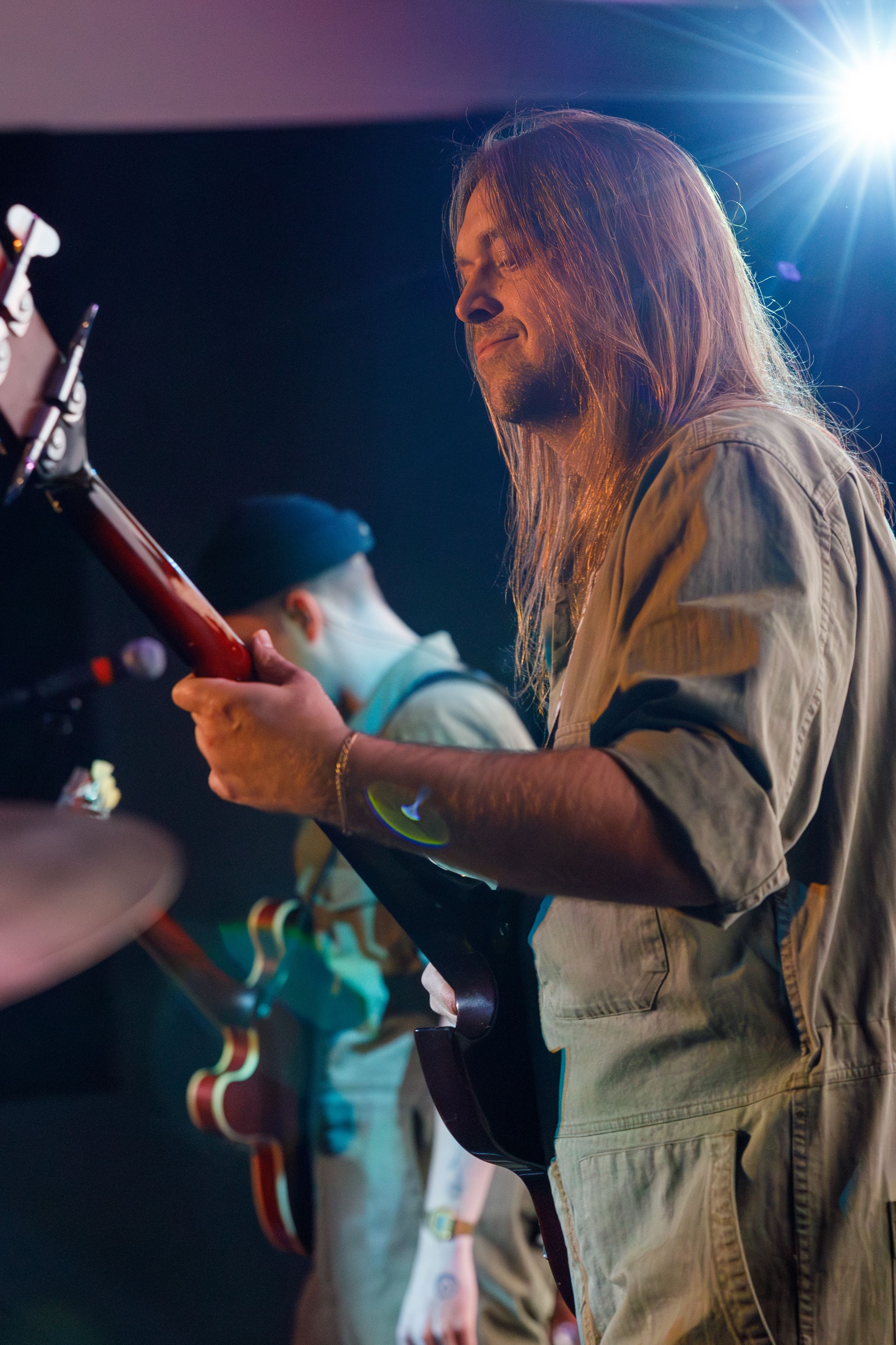 Musician performing on stage with long hair and denim shirt illuminated by bright stage lighting during live performance.