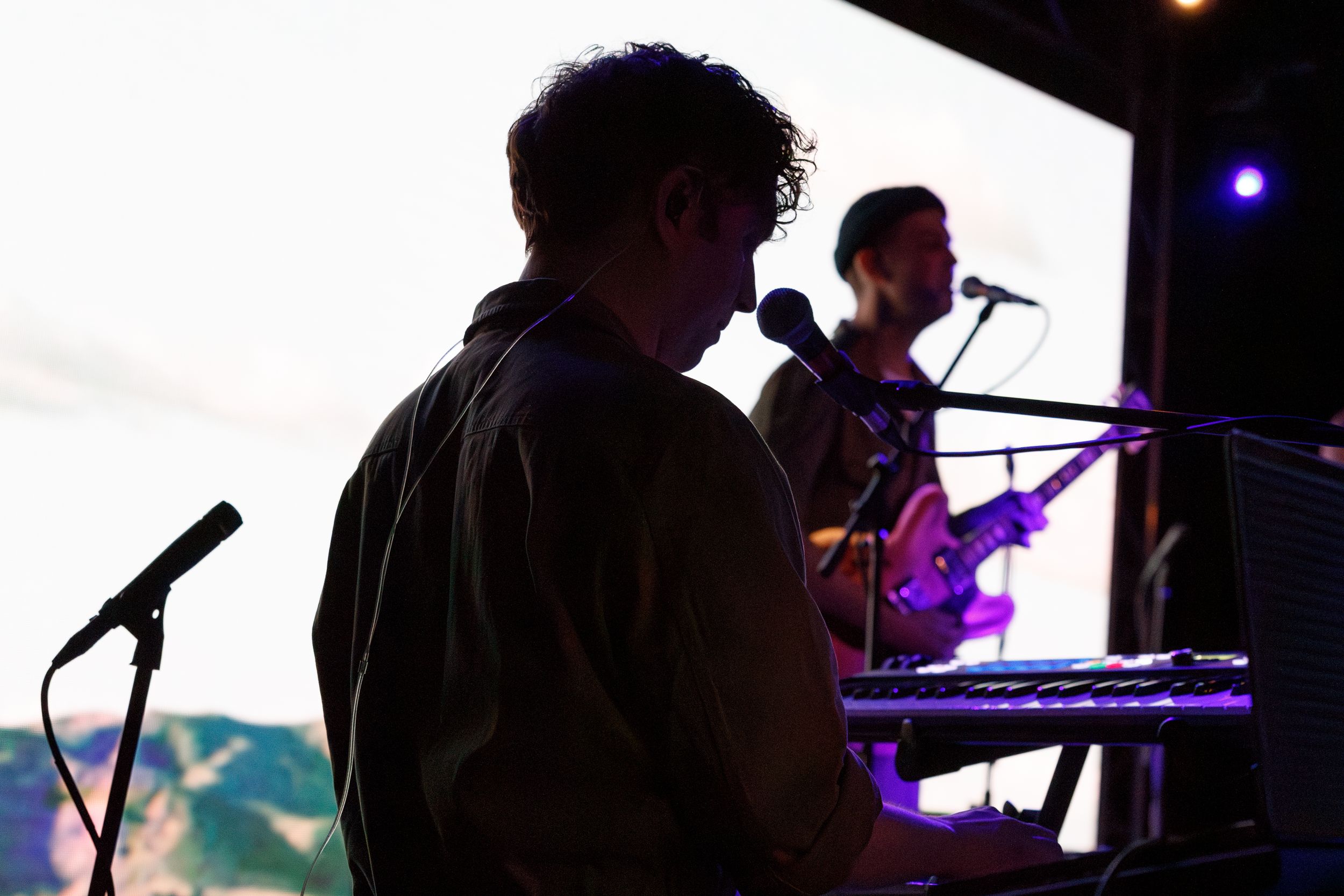 Silhouetted musicians perform on stage with keyboards and microphones against a backlit backdrop at dusk.