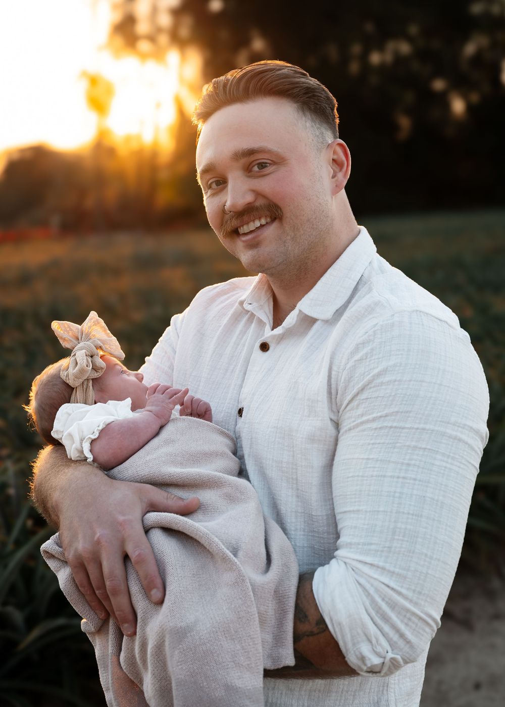 Person in a white shirt holds a newborn baby during a golden sunset photo session outdoors.