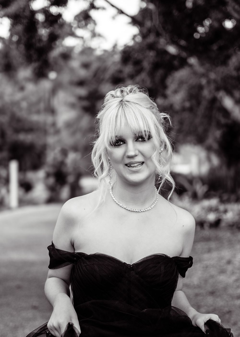 A candid black and white outdoor portrait featuring an off-shoulder black gown and delicate pearl necklace.