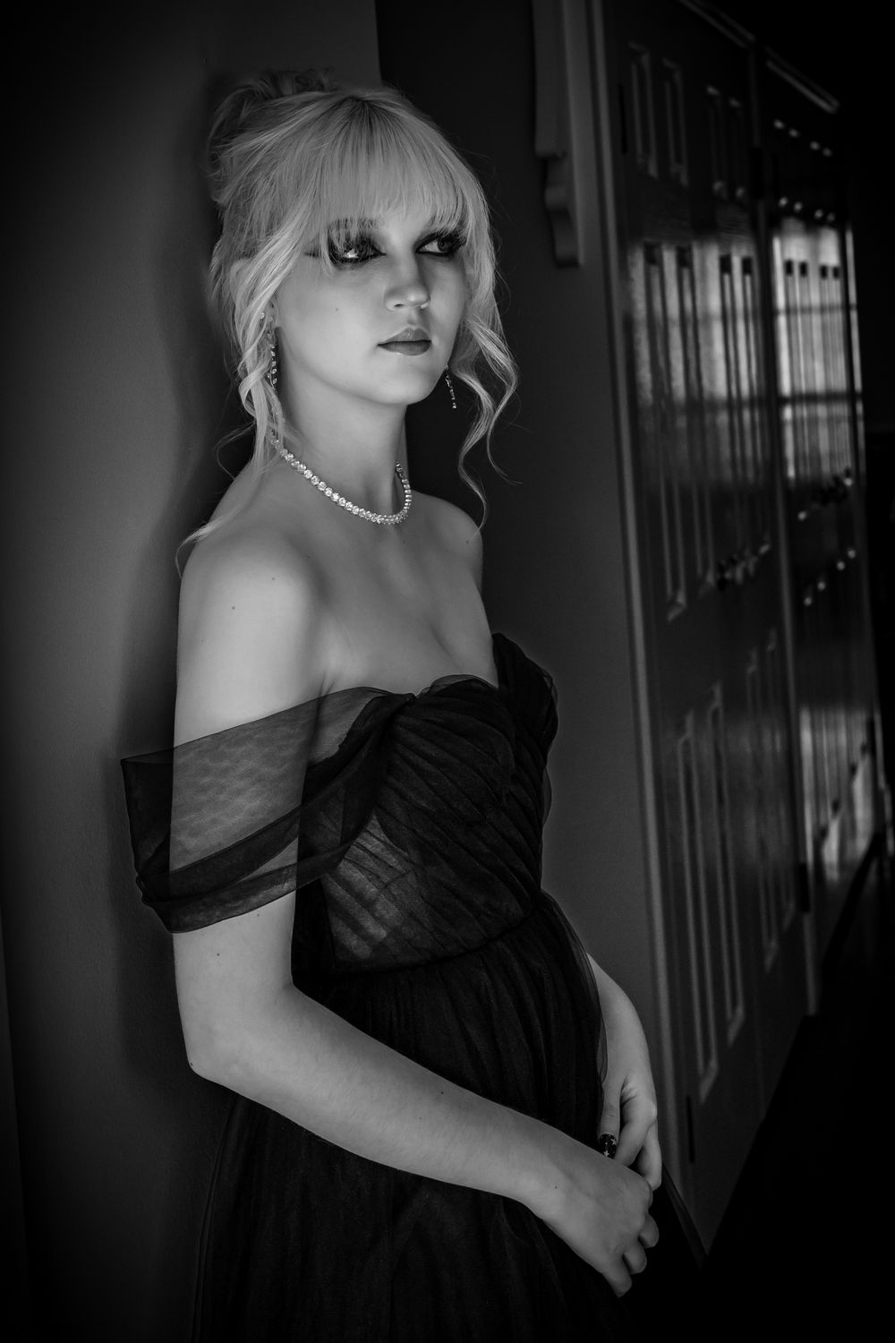 Classic black and white portrait showing an elegant off-shoulder black dress and pearl necklace in a moody setting.