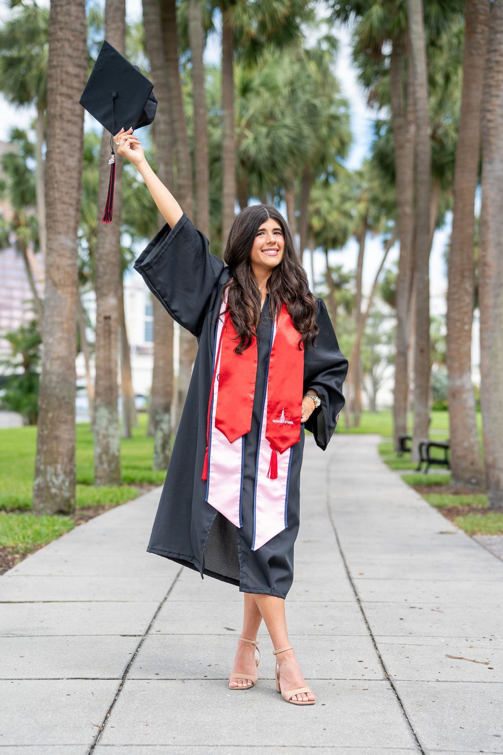 Graduate in black cap and gown with red stole celebrating on palm tree-lined walkway on sunny day.