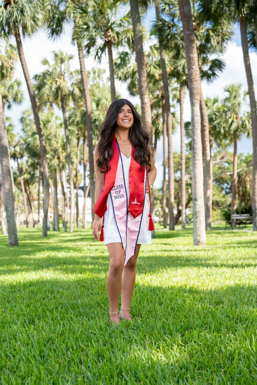 A person in graduation regalia stands smiling on green grass with tall palm trees in the background.
